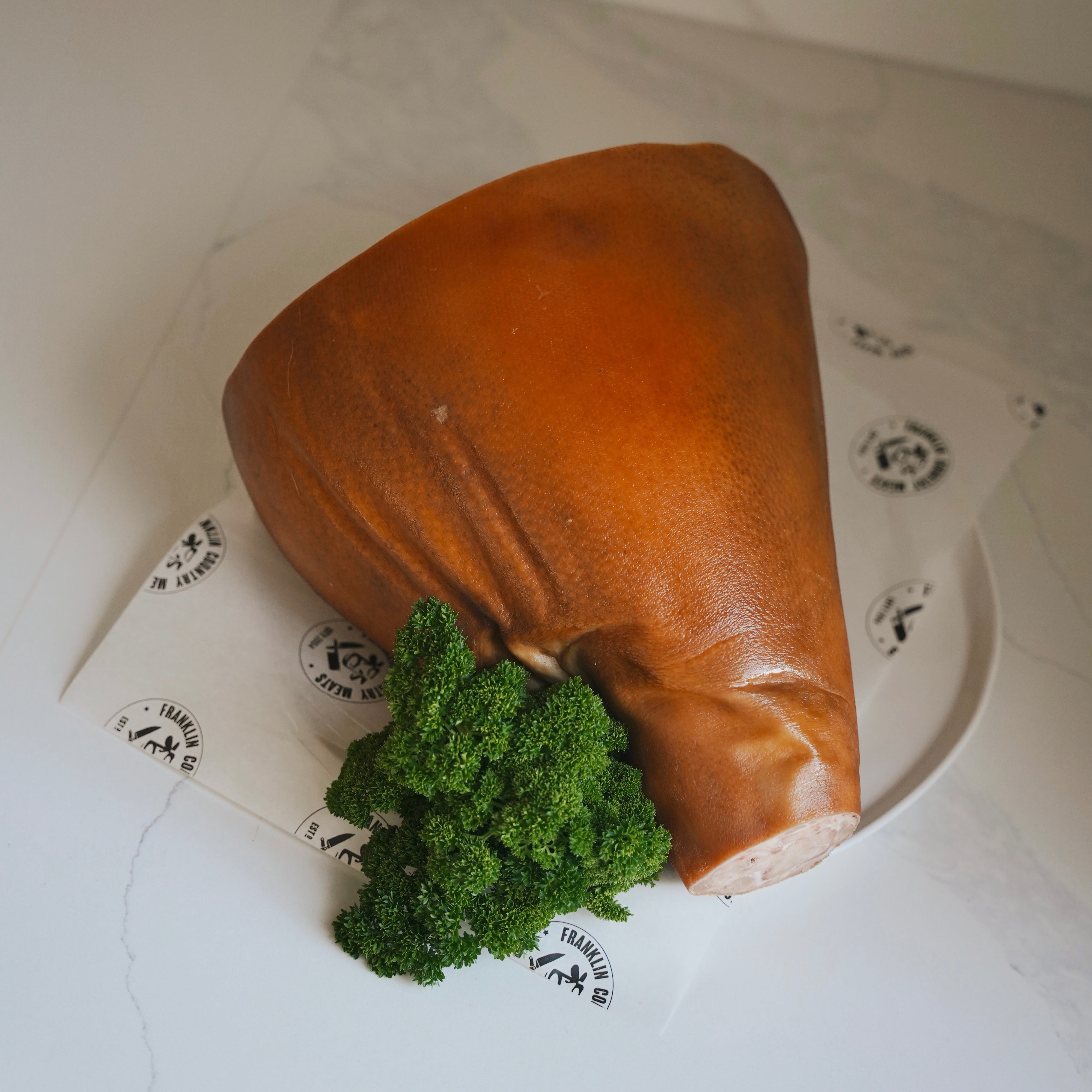 Raw ham back view on a white plate with green herbs and branded paper.